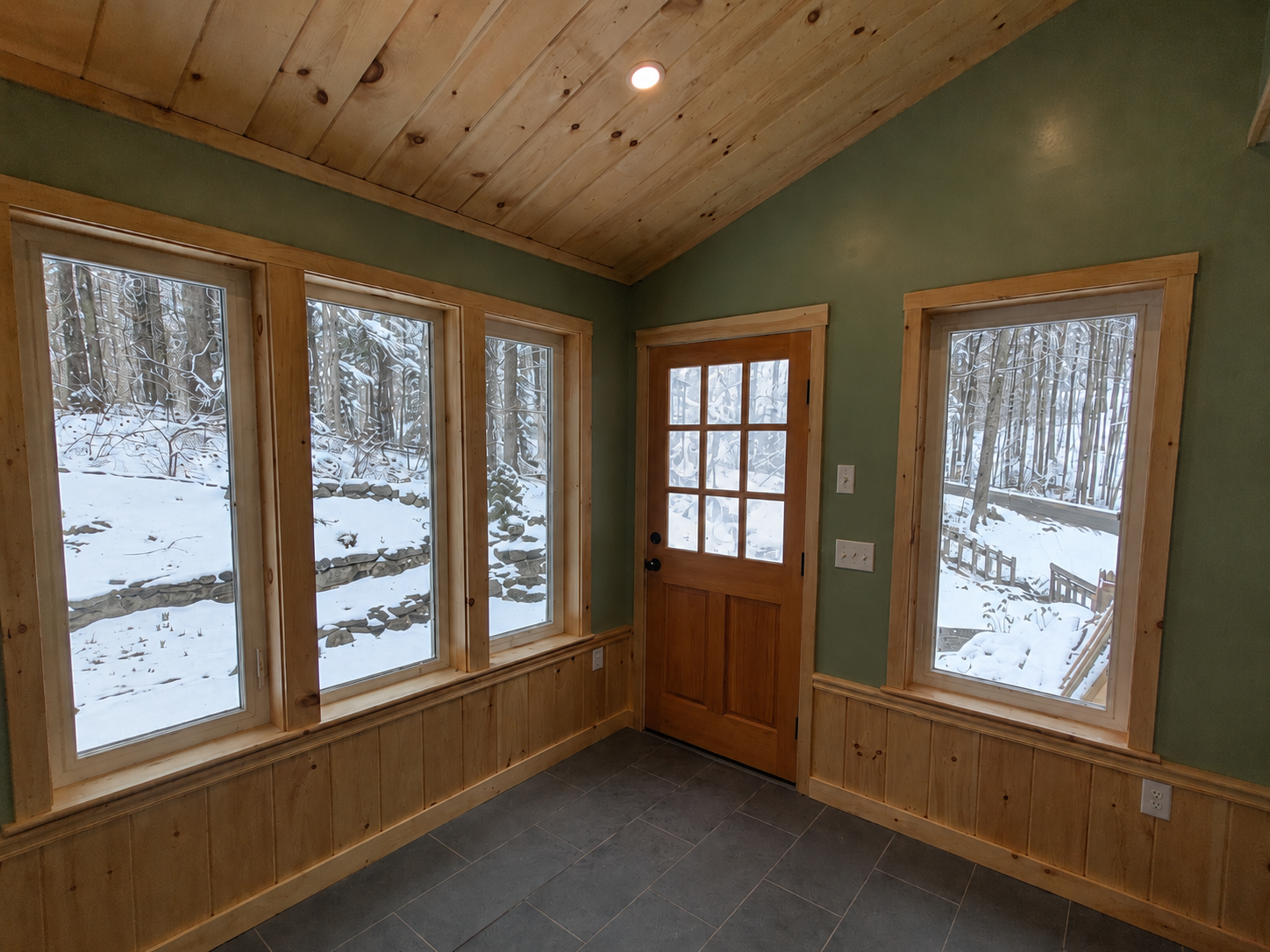 A finished mudroom with pine ceiling, sage green walls, and large windows looking into a snowy woodland